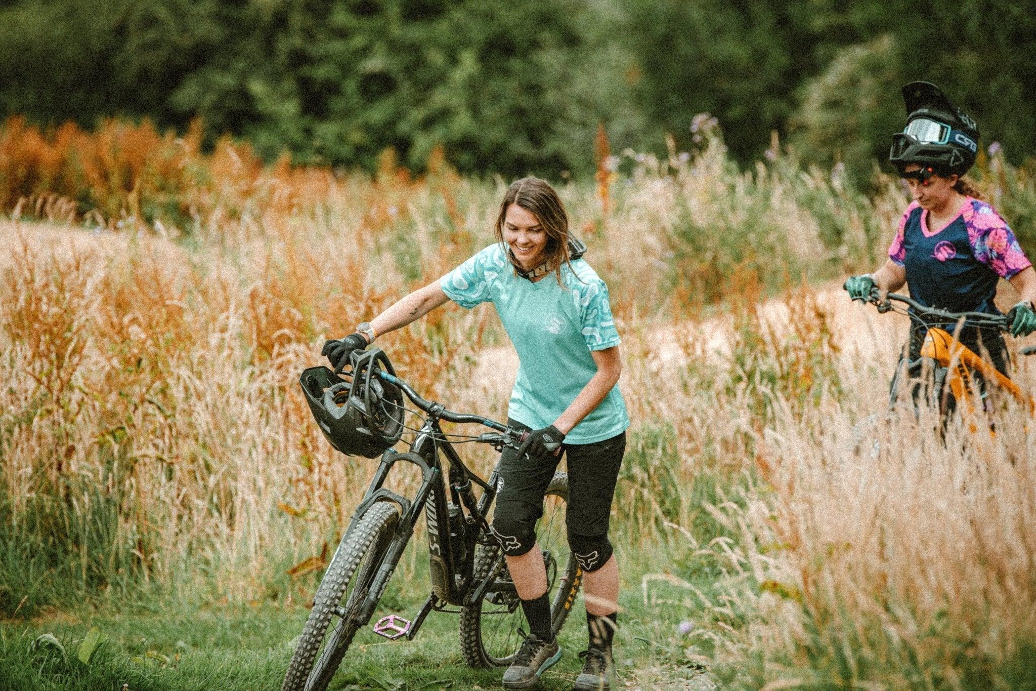 Two women with mountain bikes in a grassy trail area, wearing casual MTB jerseys and protective gear