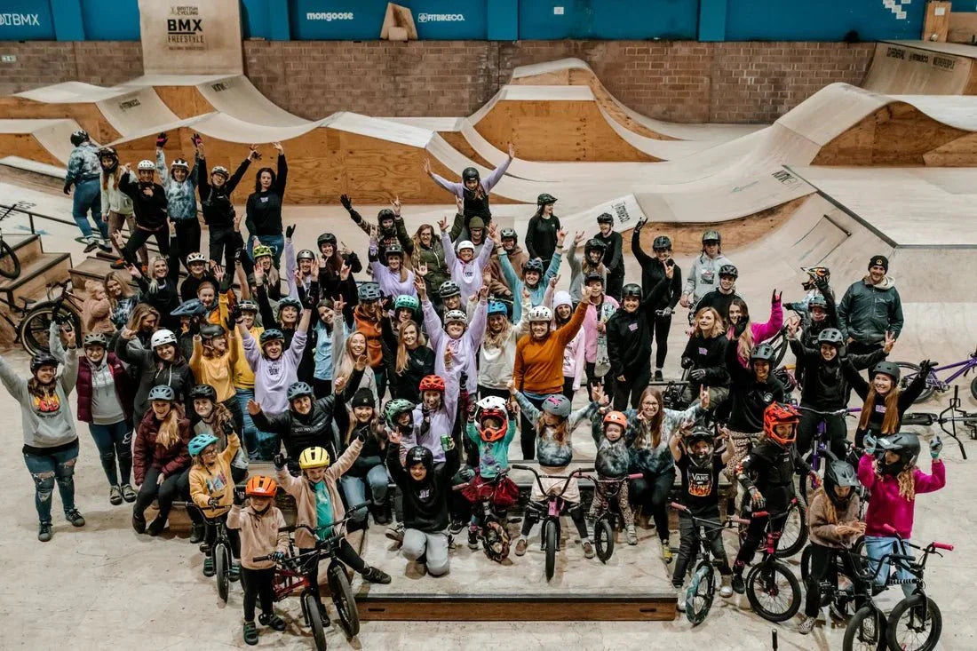 Large group of women and girls wearing helmets at an indoor BMX skatepark, posing with bikes and ramps