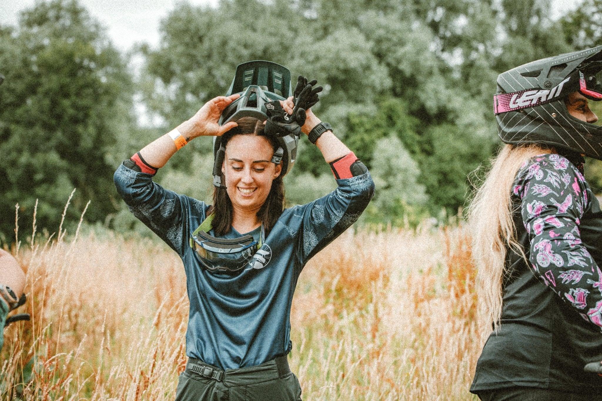 Two women in mountain bike apparel outdoors, one smiling and holding a helmet above her head, the other wearing a helmet