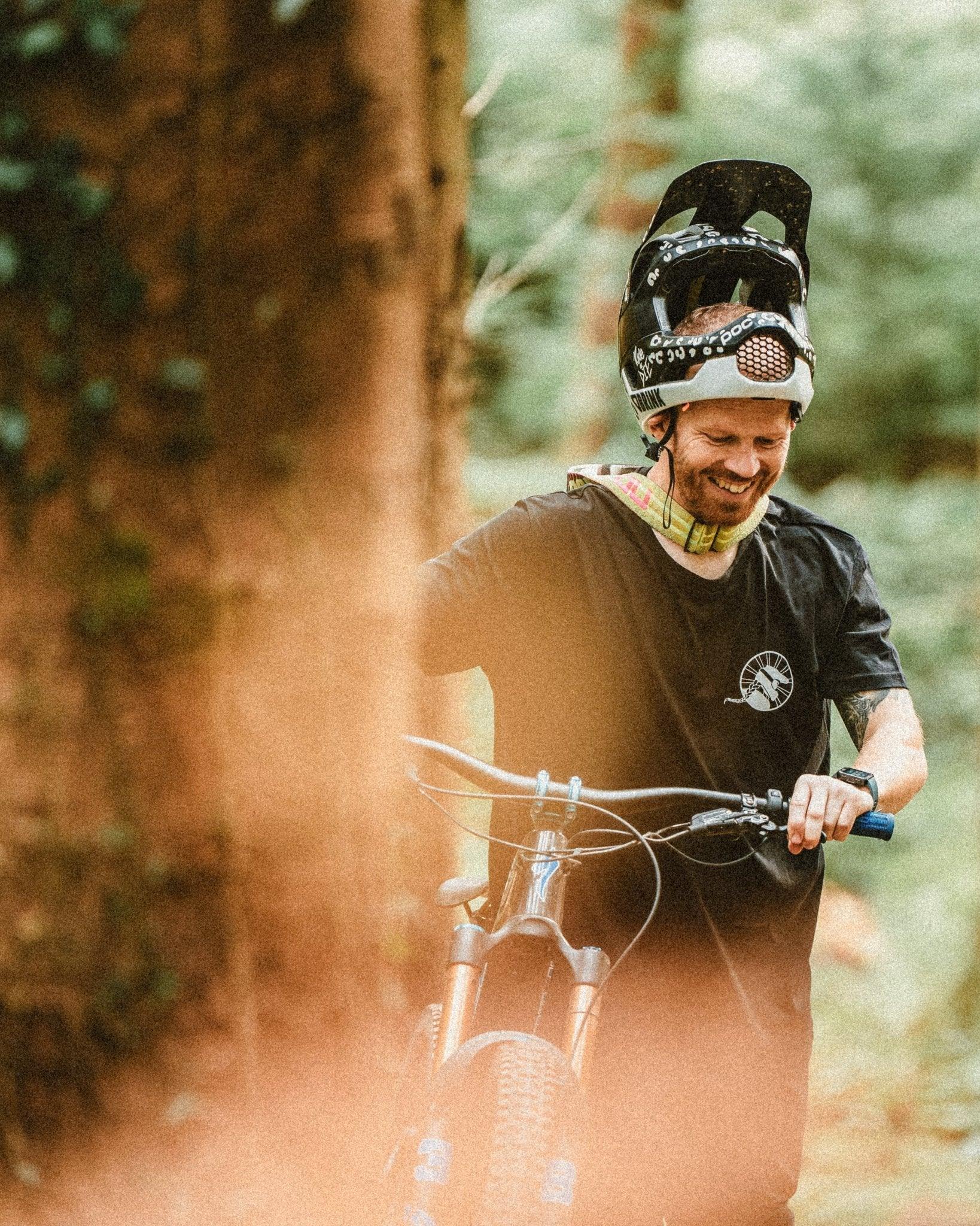 Smiling man wearing mountain bike helmet and black Unbound Collective t-shirt with MTB bike in forest