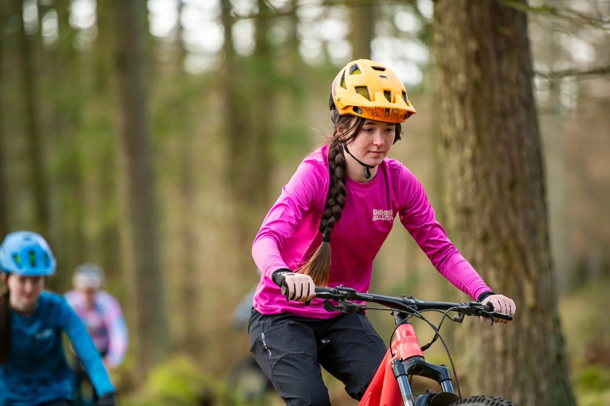 Woman mountain biking in forest wearing bright pink Unbound Collective jersey, yellow helmet, and black pants