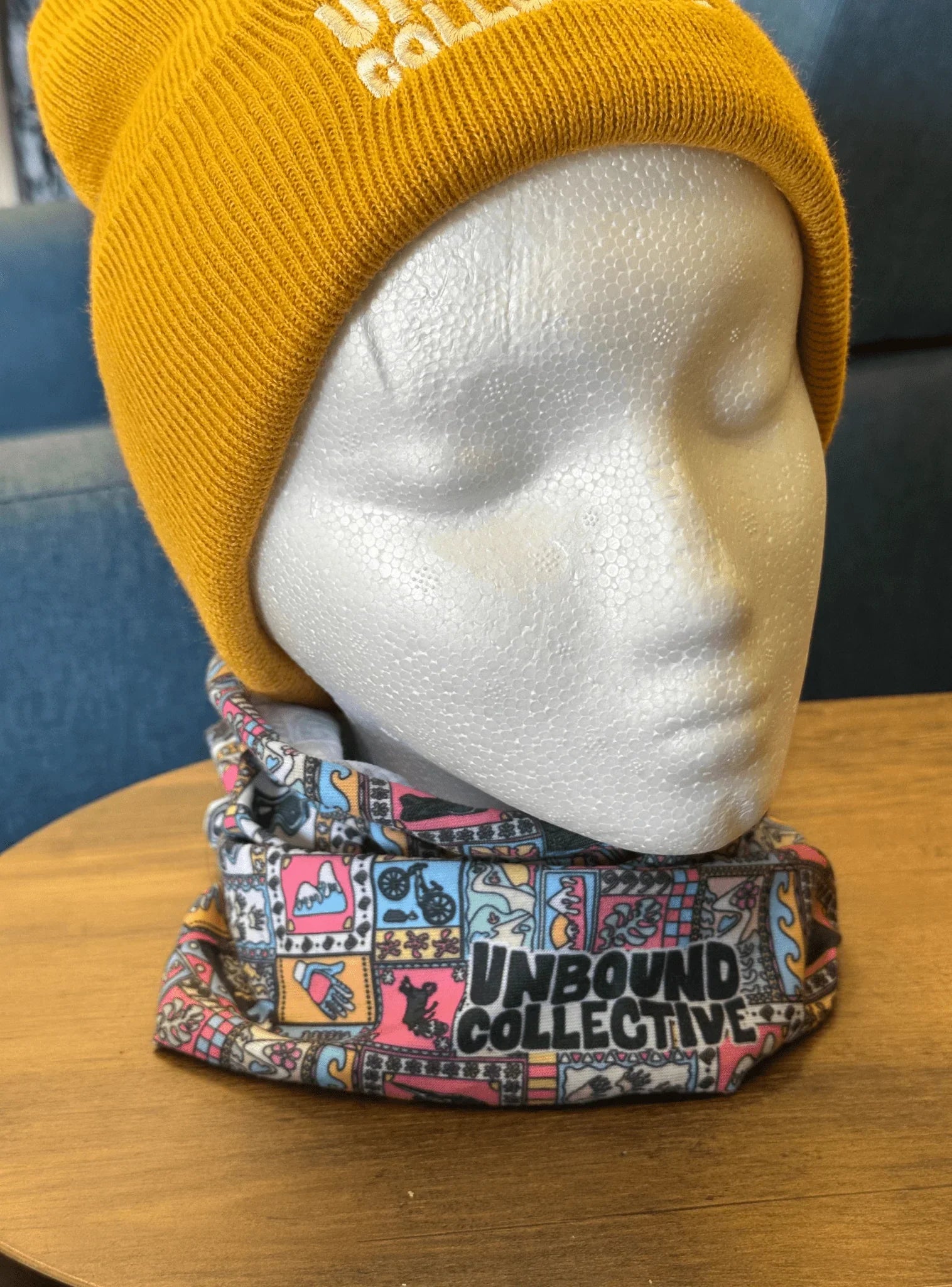 White mannequin head wearing a mustard yellow beanie and a colorful patterned neck tube with Unbound Collective logo