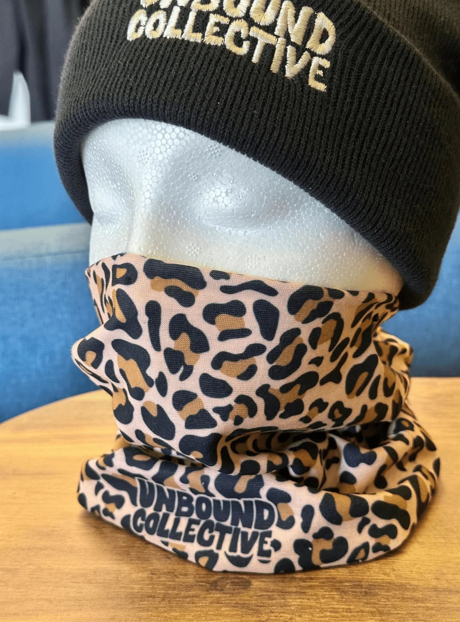 Styrofoam head wearing a black Unbound Collective beanie and leopard print neck tube with Unbound Collective logo on wooden table