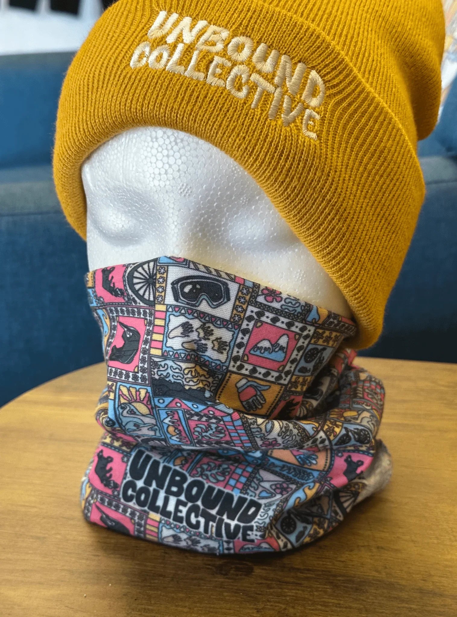 White mannequin head wearing a mustard-yellow knit beanie with Unbound Collective logo and a colorful patterned neck tube with Unbound Collective branding