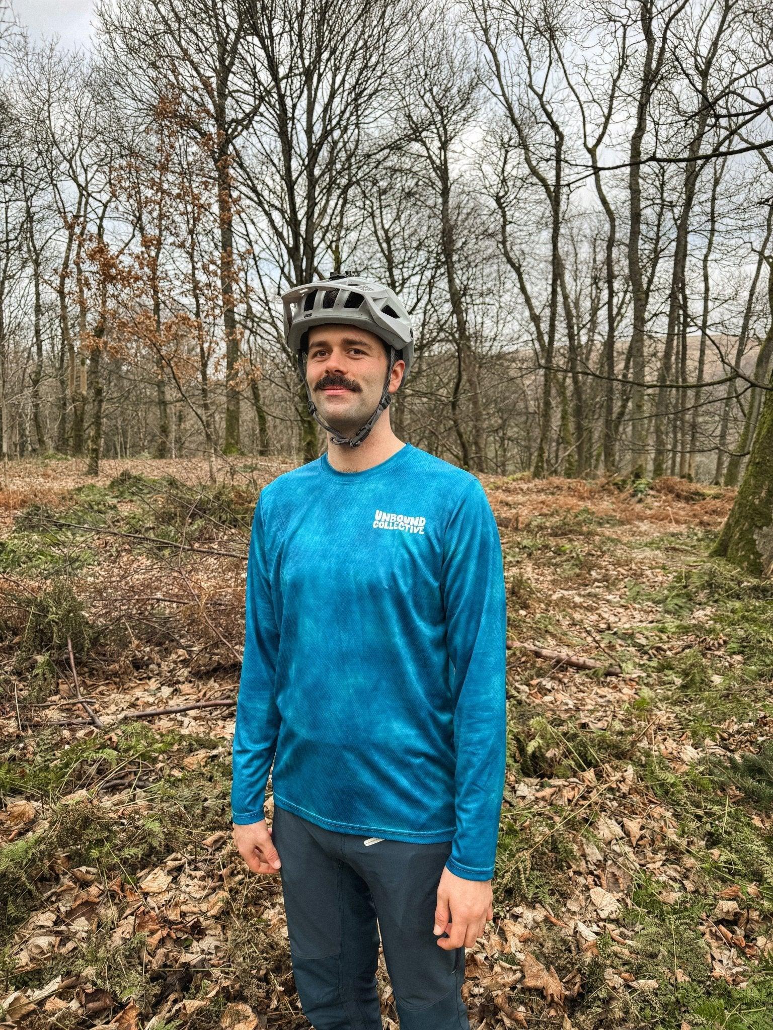 Man wearing blue Unbound Collective mountain bike jersey and gray helmet in leaf-covered forest