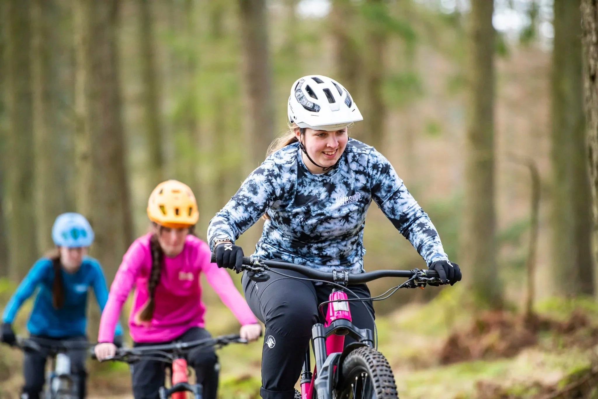 Woman wearing white helmet and smoke pattern MTB jersey mountain biking in forest with two others