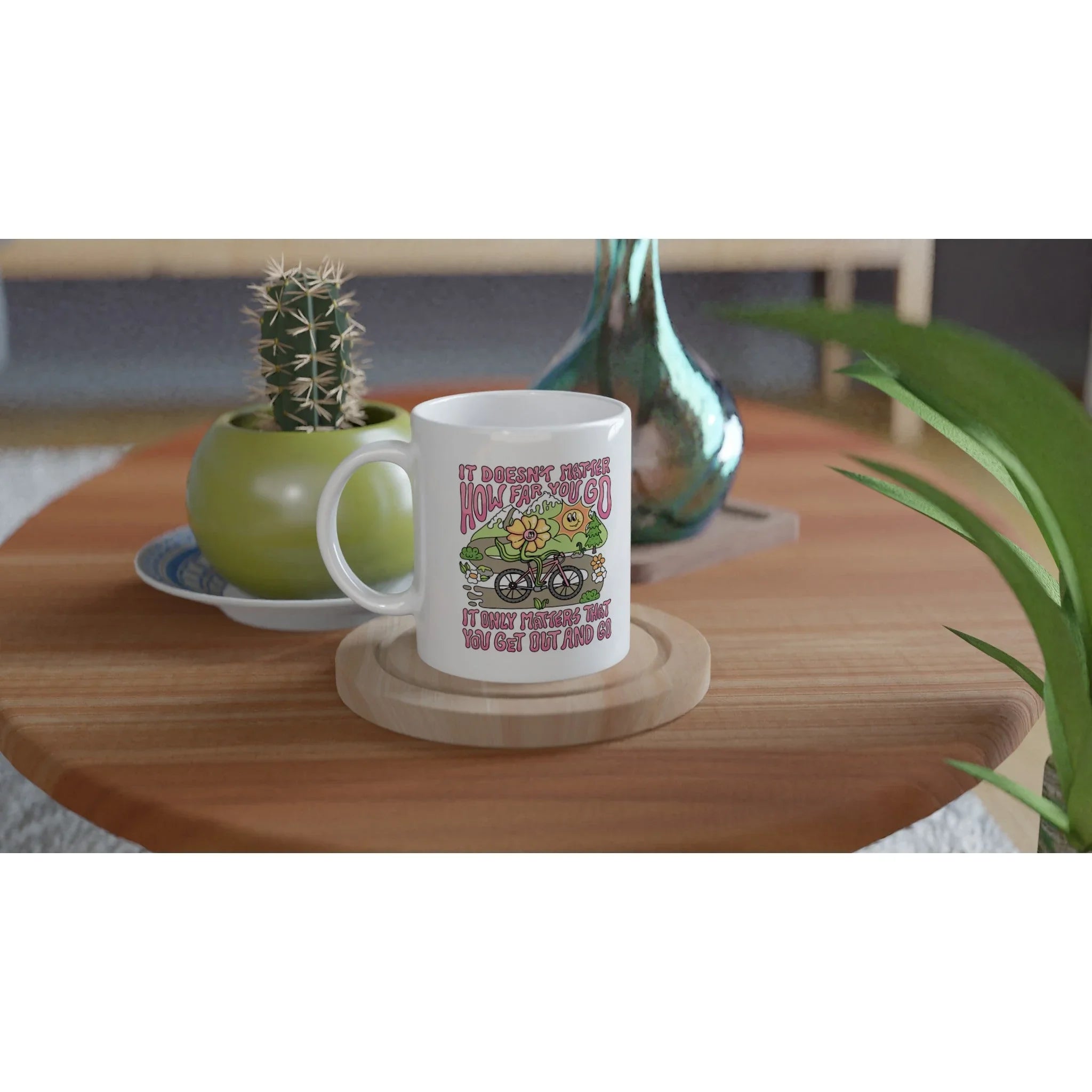 White ceramic mug with colorful motivational cycling design on wooden table with green potted cactus and glass vase