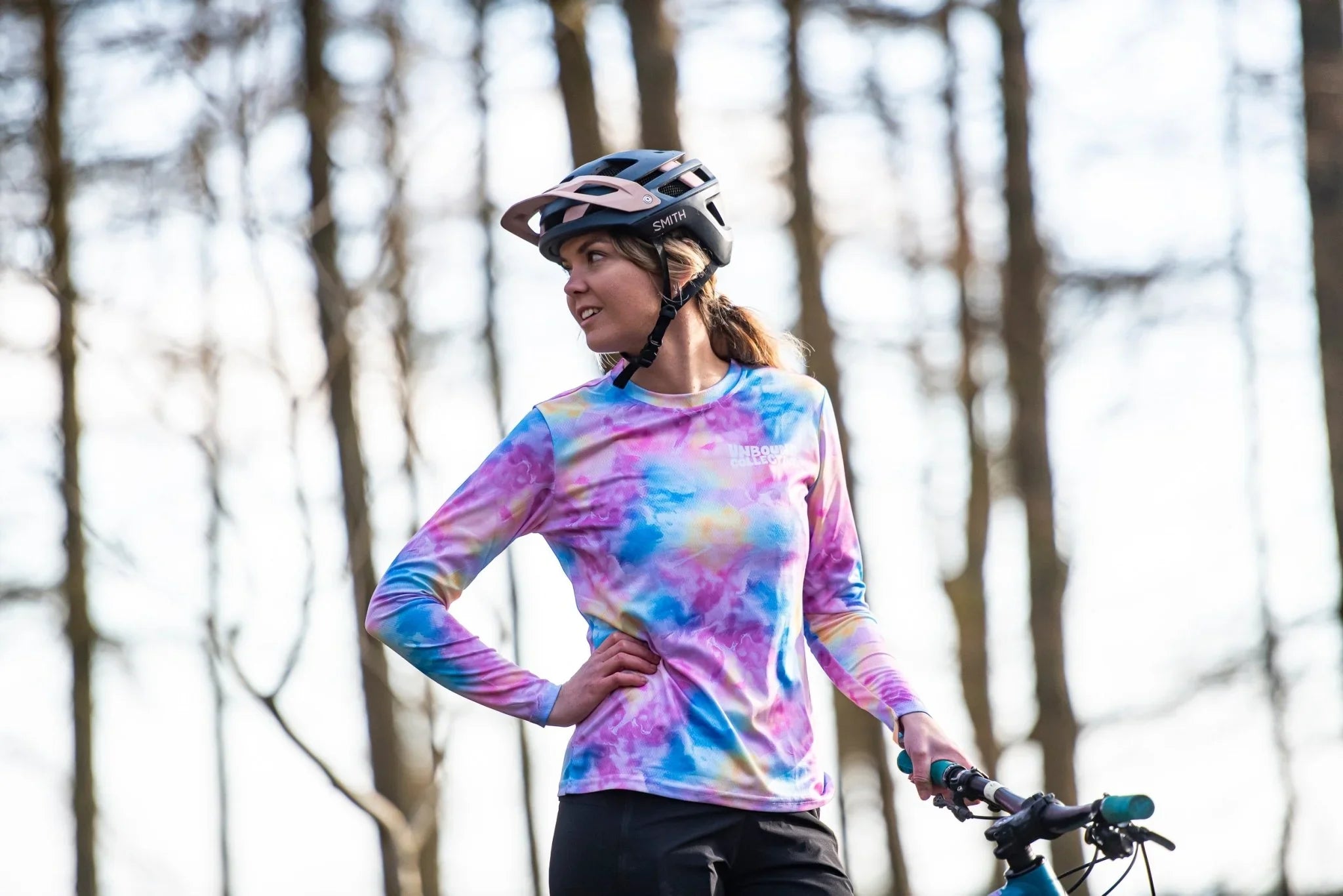 Woman wearing tie-dye long sleeve MTB jersey and helmet standing with mountain bike in forest