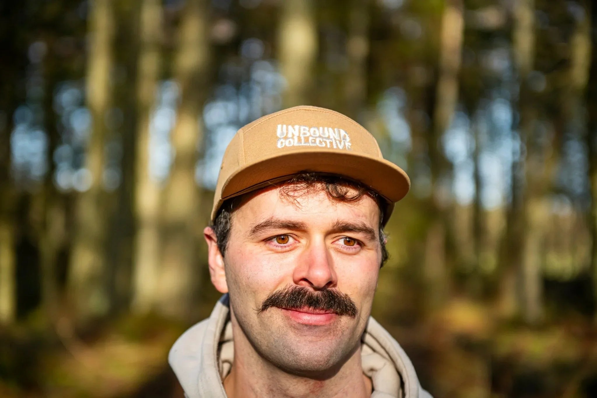 Man wearing beige Unbound Collective 5-panel hat outdoors in sunlit forest background