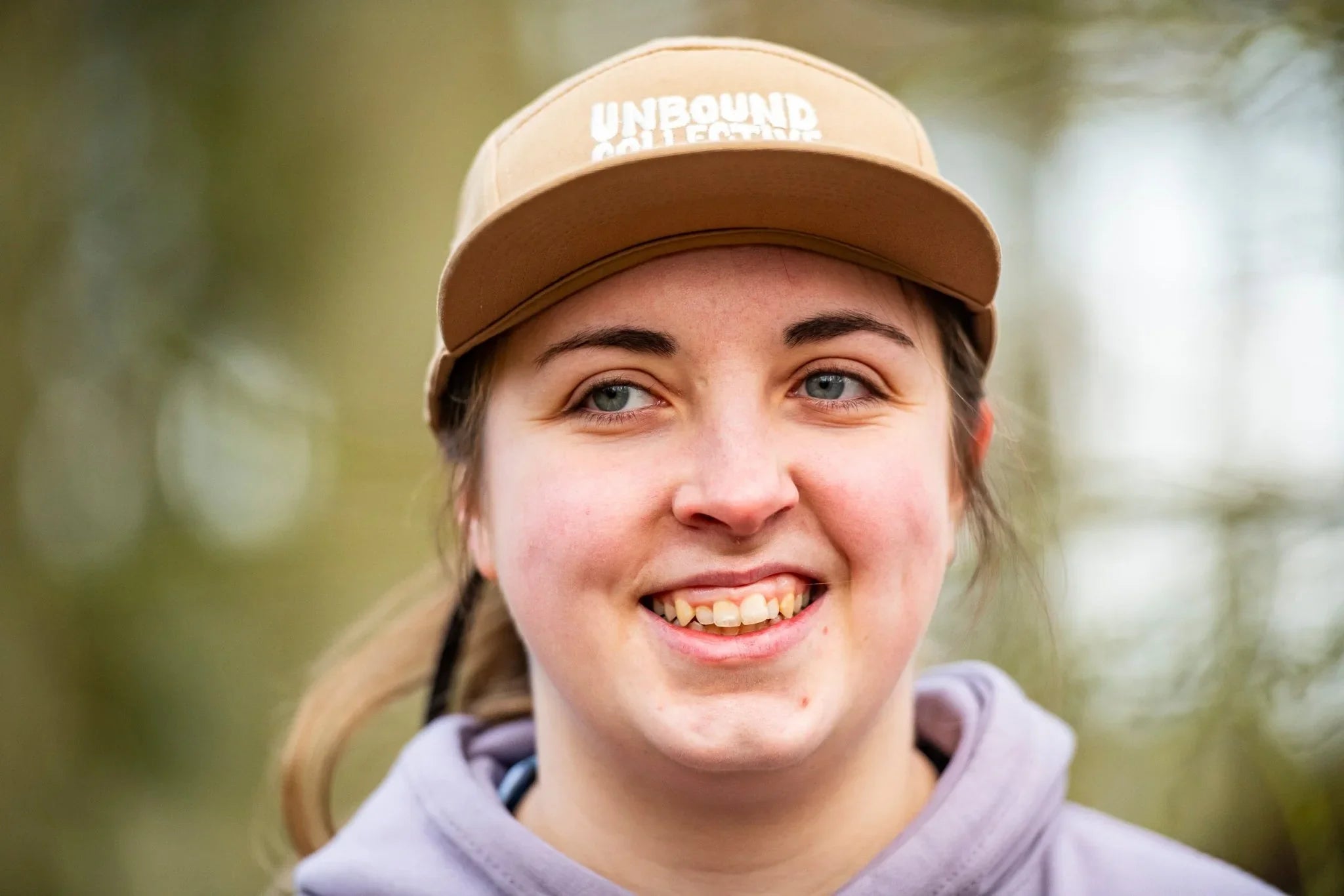 Close-up of woman wearing a tan Unbound Collective 5-panel hat outdoors, smiling in natural setting