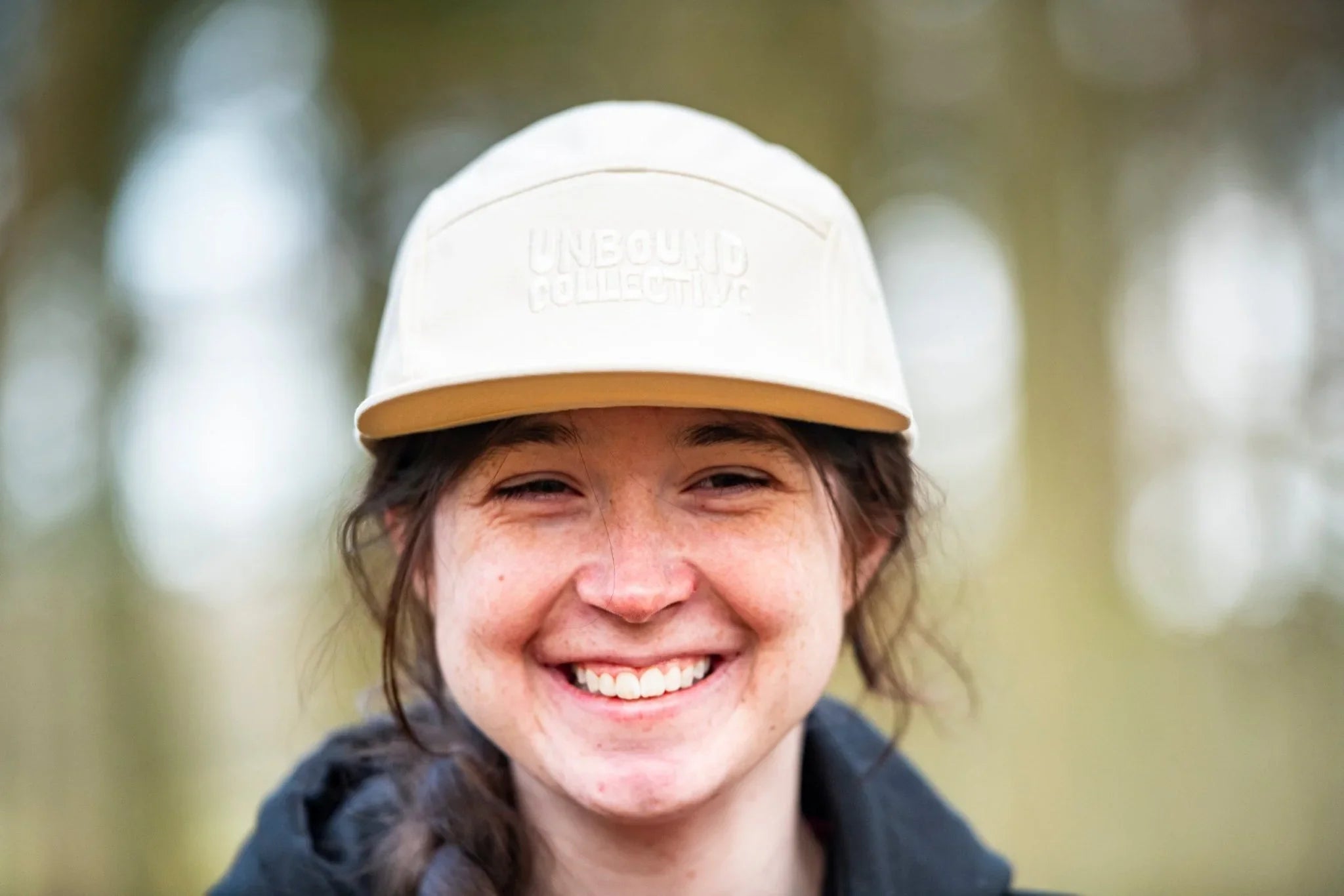 Smiling woman wearing a white Unbound Collective 5-panel hat in an outdoor forest setting