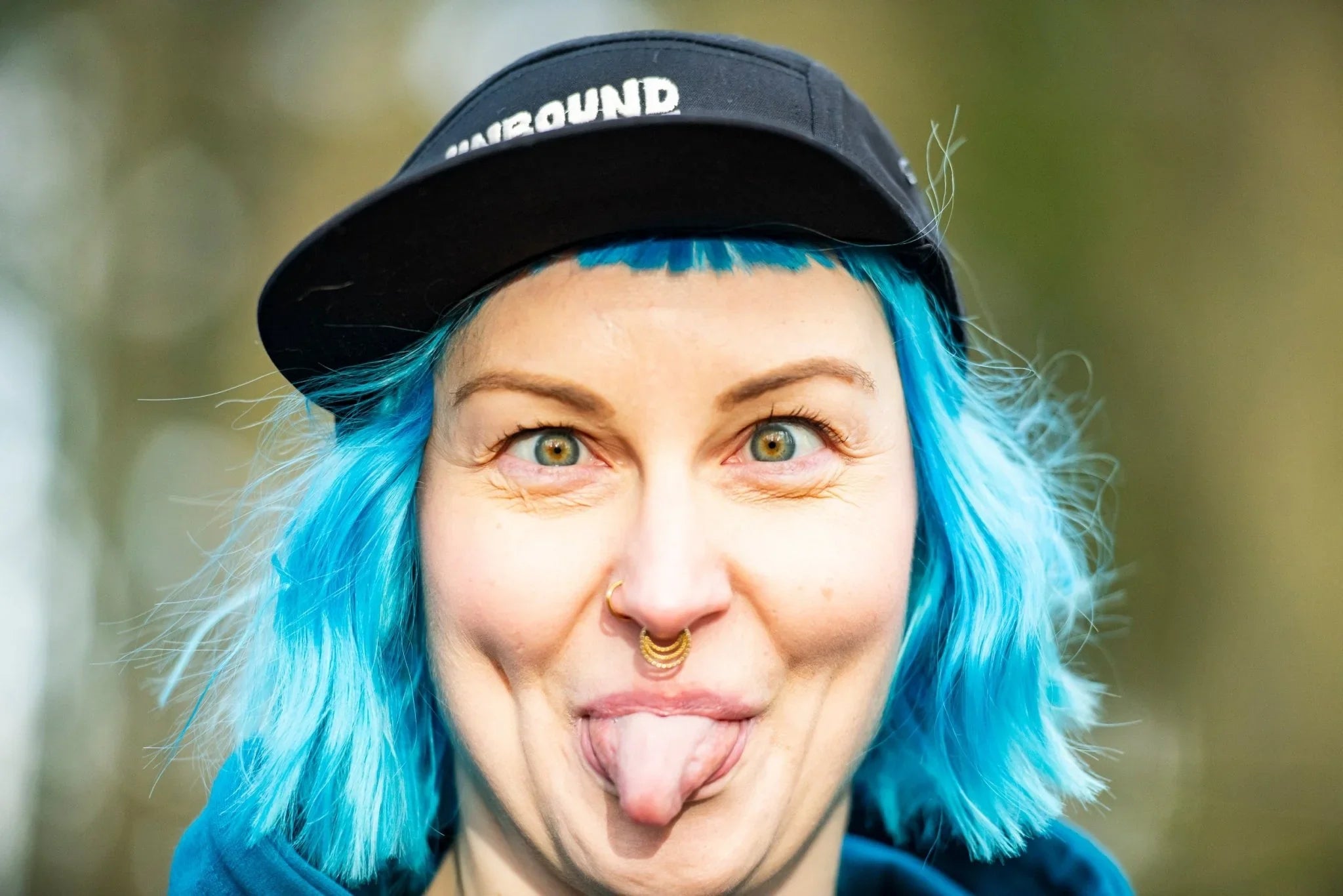 Person with bright blue hair wearing black Unbound cap sticking out tongue outdoors