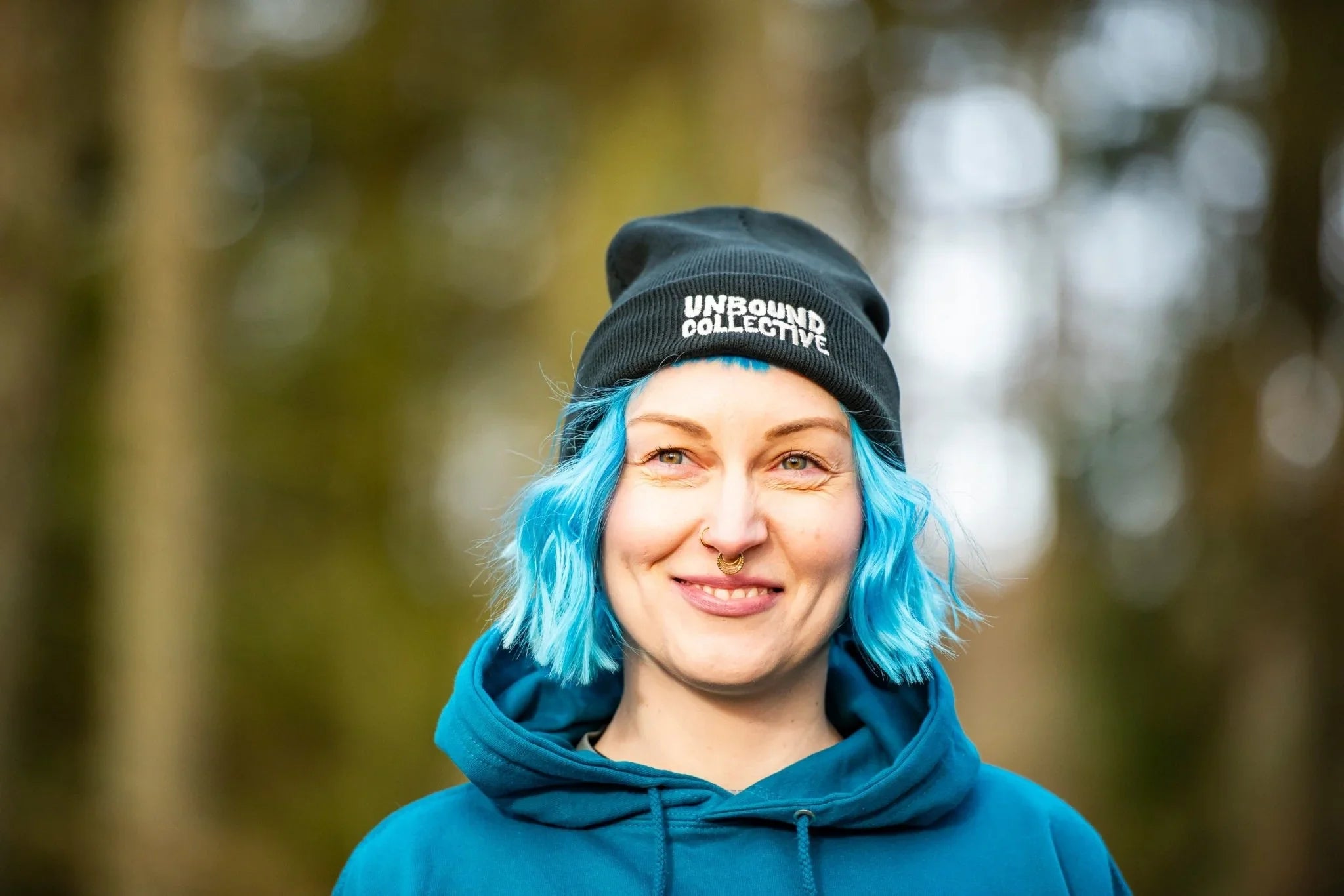 Smiling woman with blue hair wearing a black Unbound Collective beanie and teal hoodie outdoors