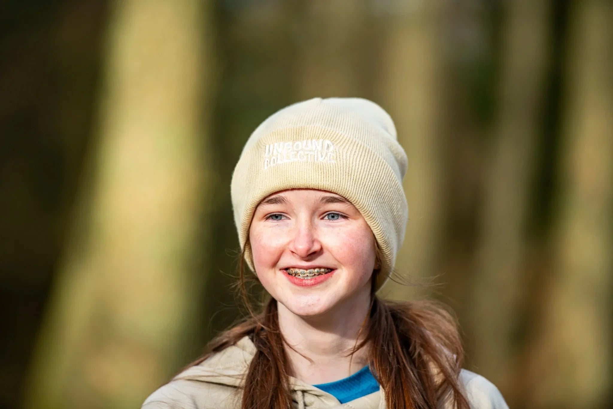 Smiling young woman outdoors wearing a beige knit beanie hat with Unbound Collective logo, casual hoodie
