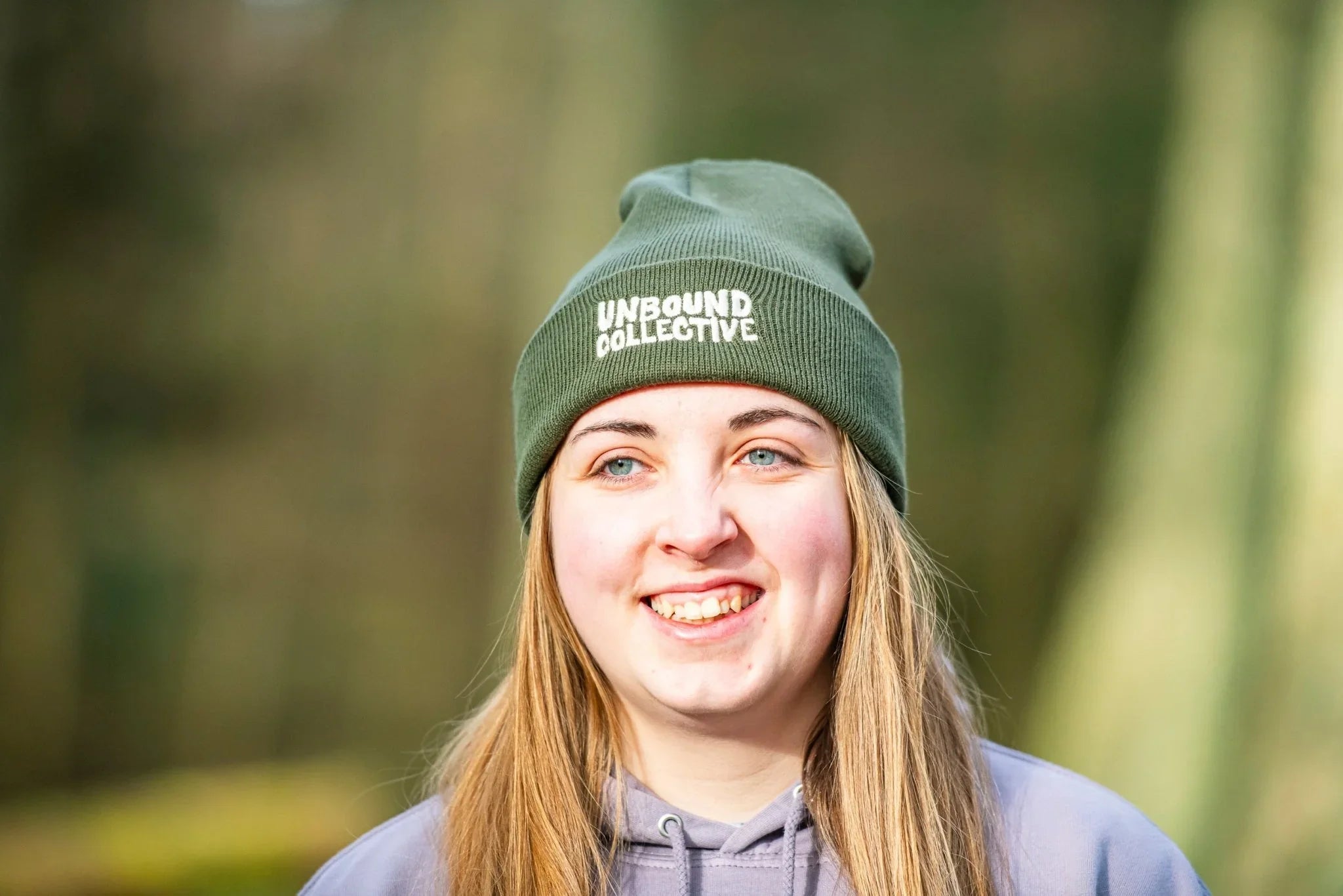 Smiling woman wearing an olive green Unbound Collective beanie and gray hoodie outdoors in natural light
