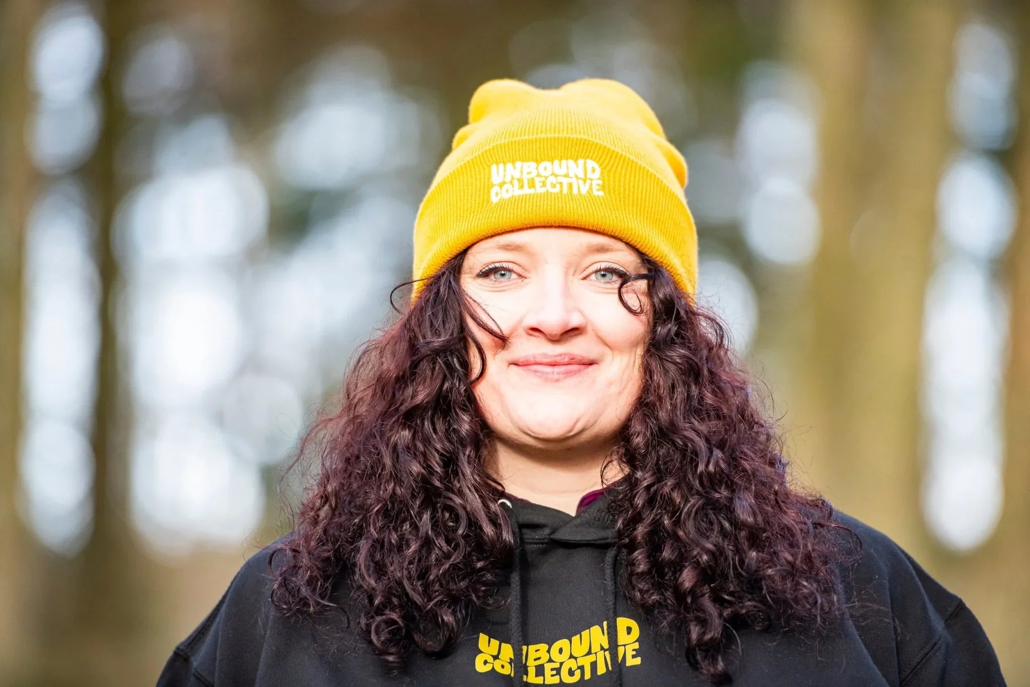 Woman wearing a yellow Unbound Collective beanie and black hoodie with outdoor blurred background