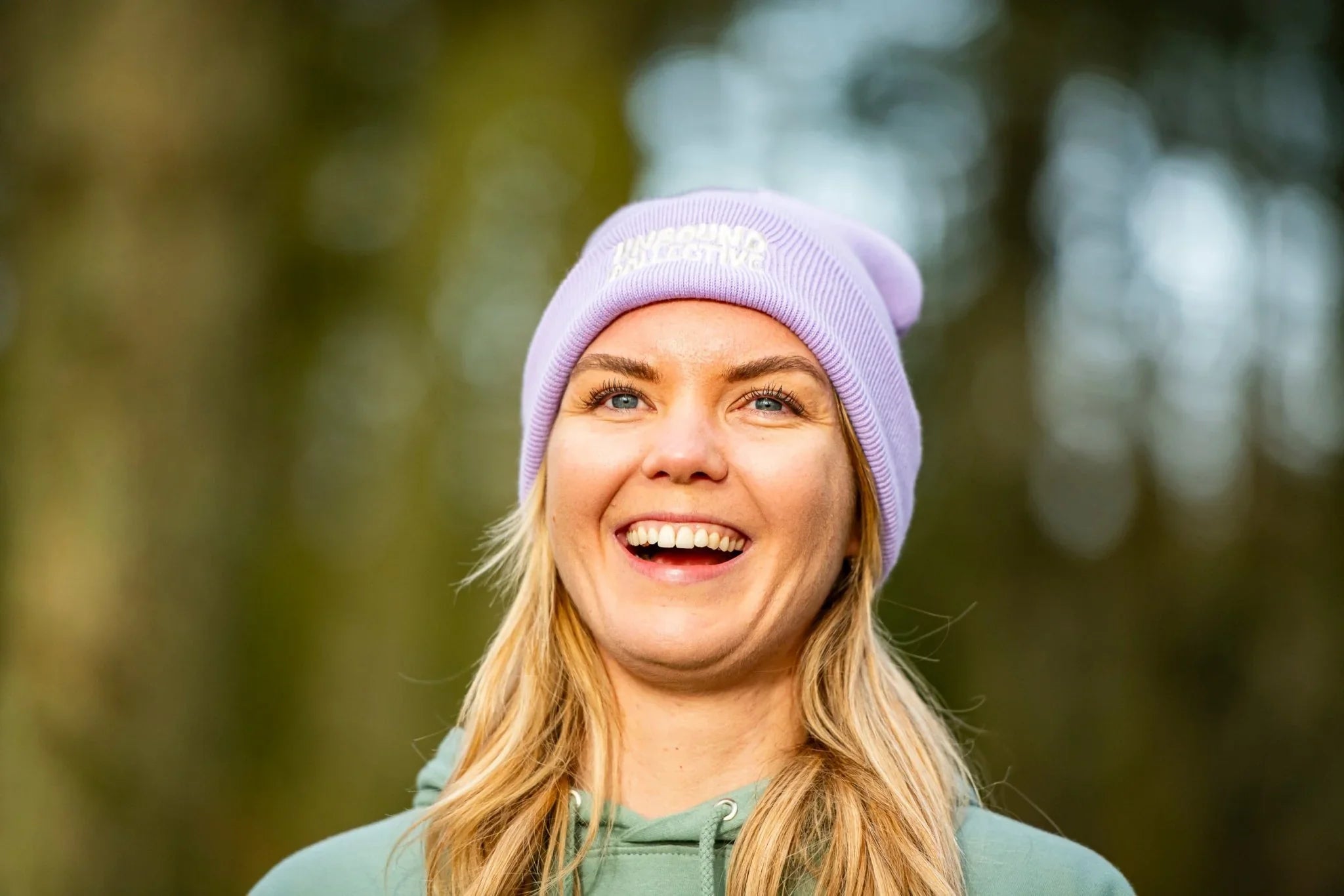 Smiling woman wearing a light purple Unbound Collective beanie hat and green hoodie outdoors