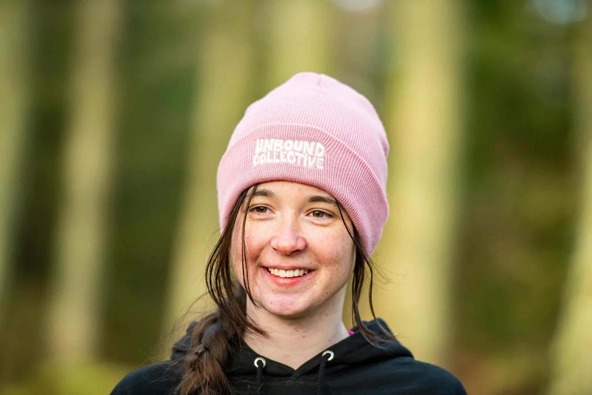 Smiling woman wearing a pink Unbound Collective beanie hat and black hoodie in an outdoor forest setting