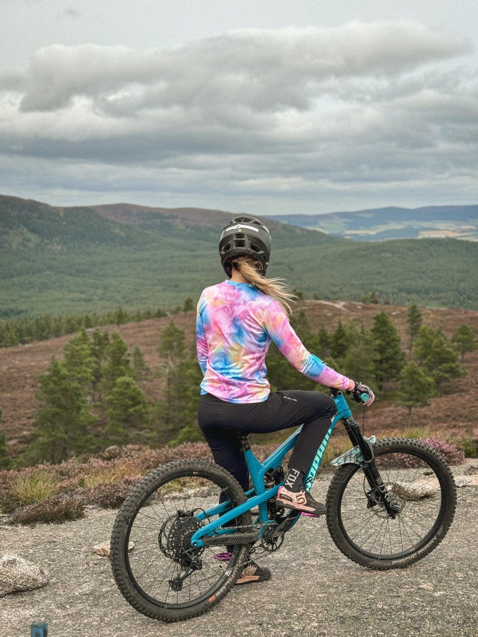 Woman in colorful MTB jersey and black pants riding blue mountain bike overlooking forested hills