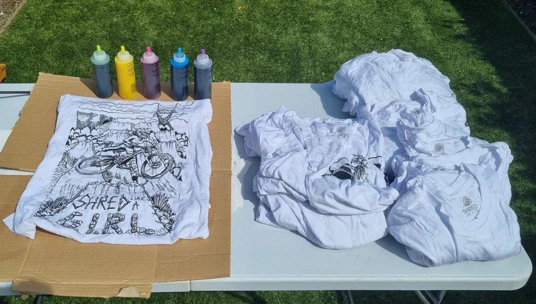 Outdoor table with white mountain bike tees, one featuring a detailed bike and 'Shred Like a Girl' design, dye bottles ready for tie-dye