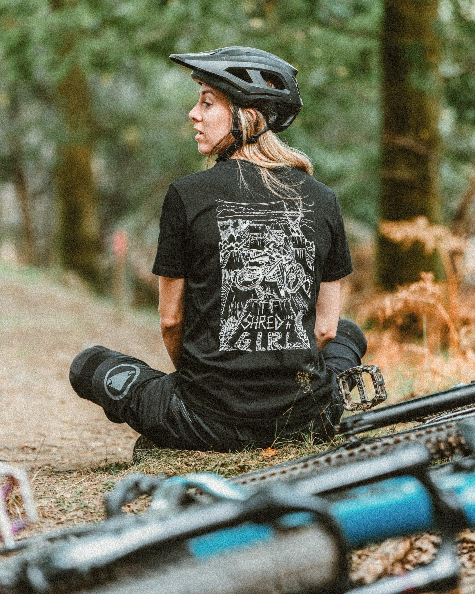 Woman in a mountain biking helmet and black tee sitting on forest trail next to a mountain bike