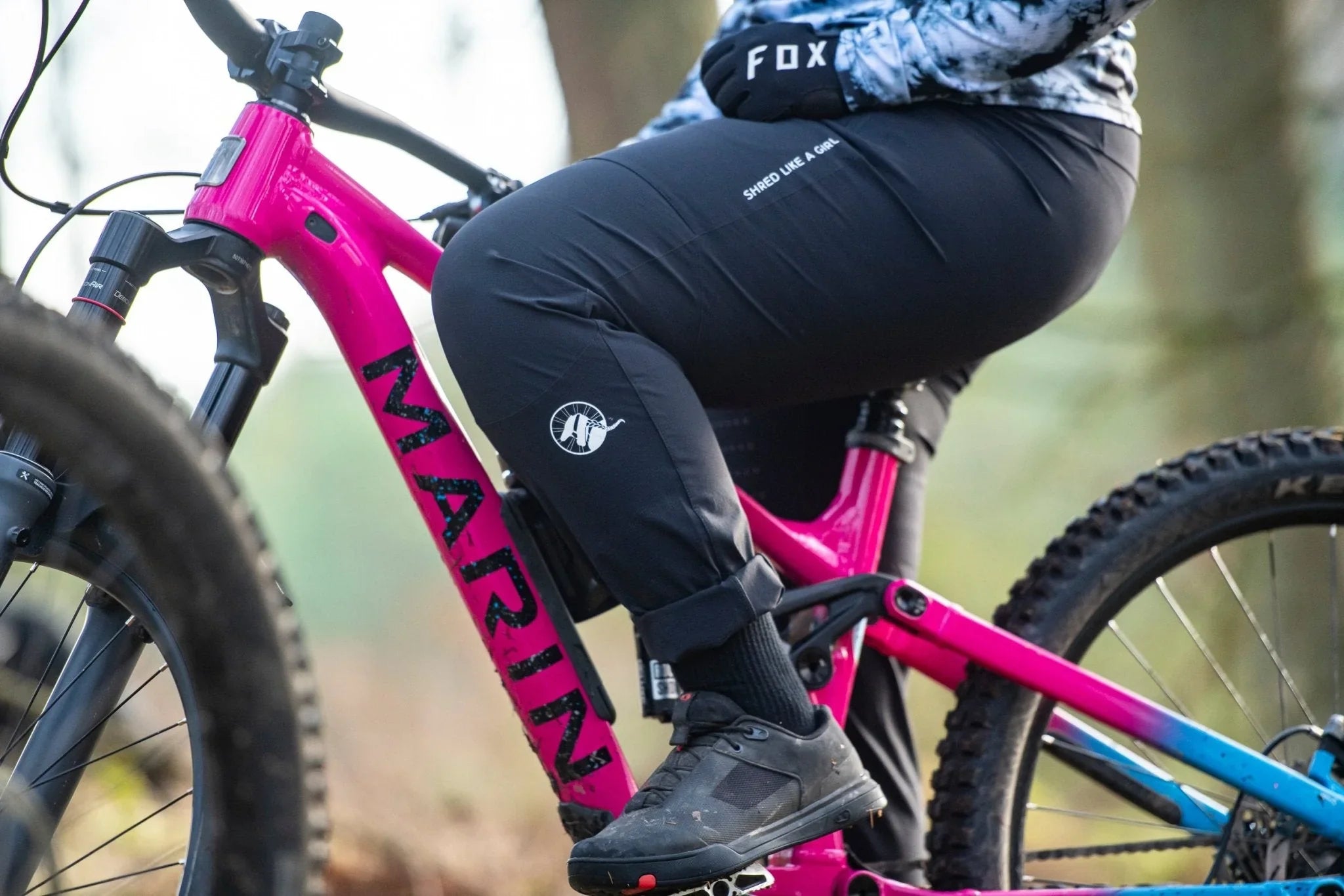 Close-up of woman wearing black MTB pants with 'Shred Like A Girl' text, sitting on pink Marin mountain bike