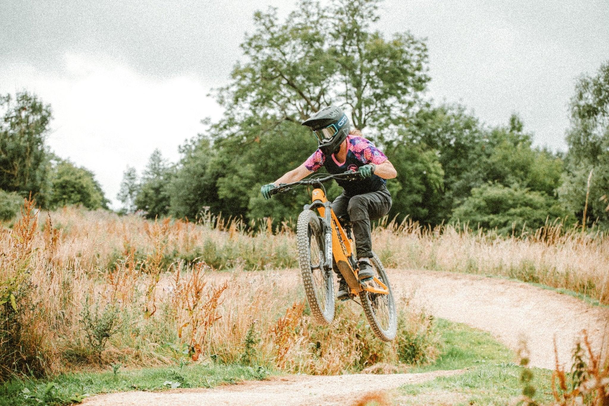 Mountain biker in full gear jumping an orange bike on a dirt trail surrounded by grass and trees