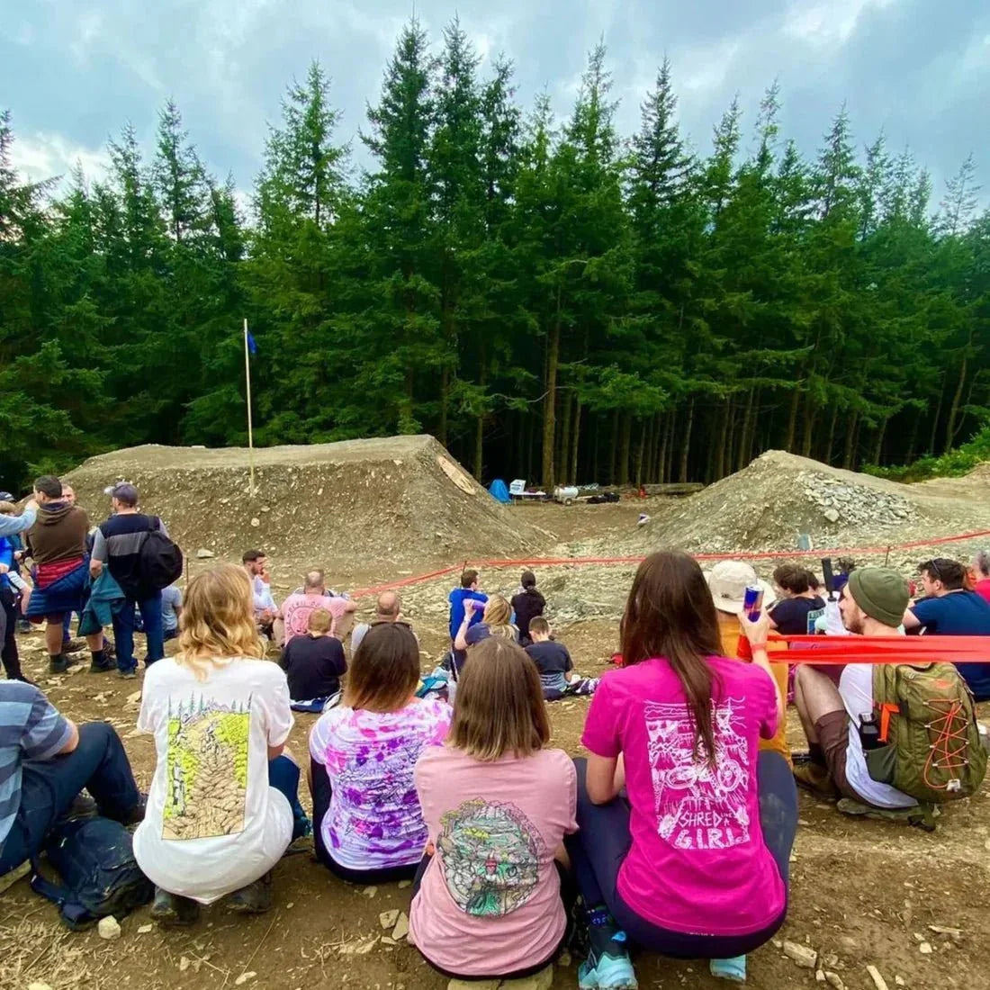 Group of spectators in colorful MTB apparel watching mountain bike dirt jumps under cloudy sky