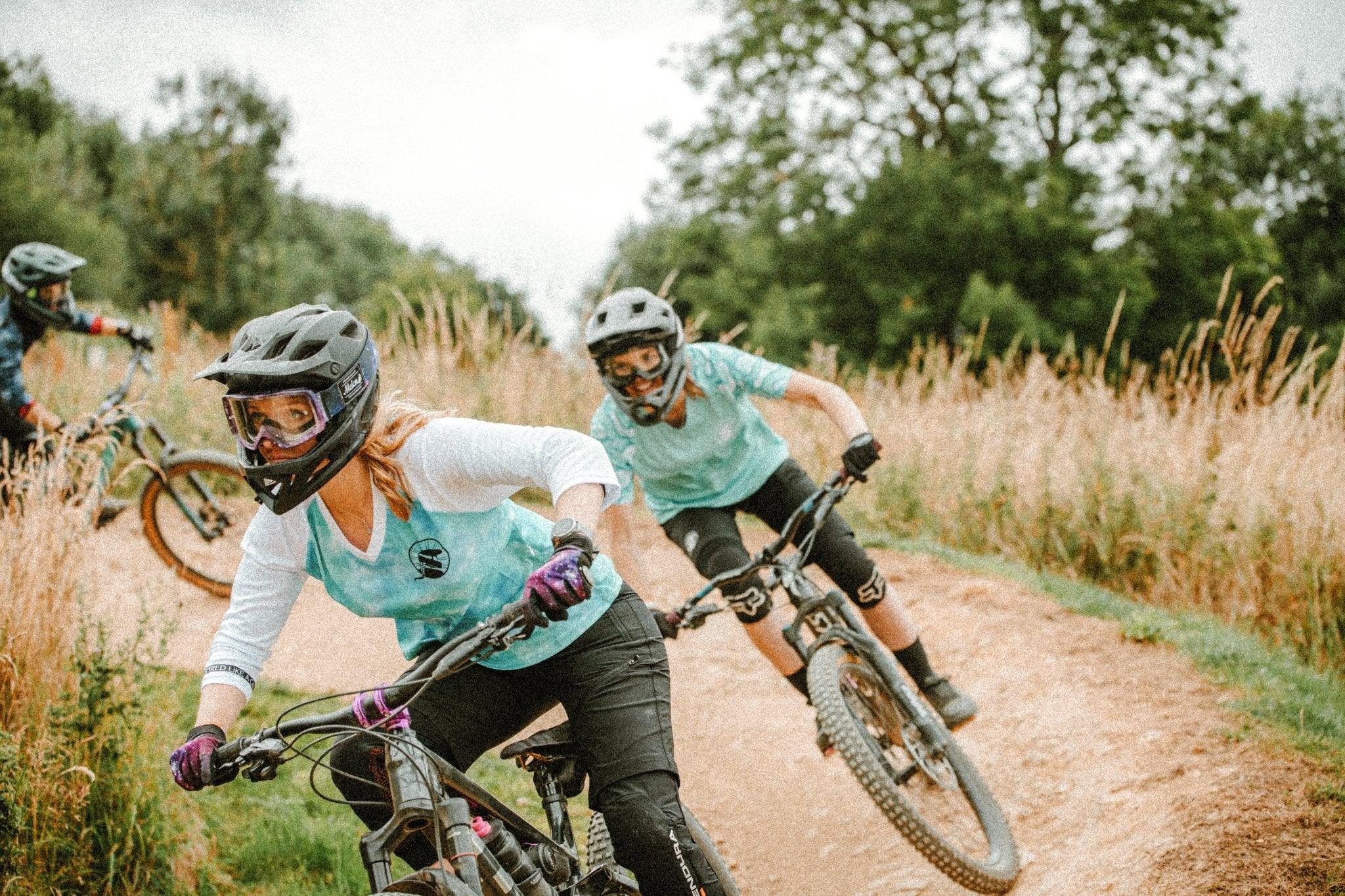 Women mountain biking on dirt trail wearing protective helmets and Unbound Collective MTB apparel