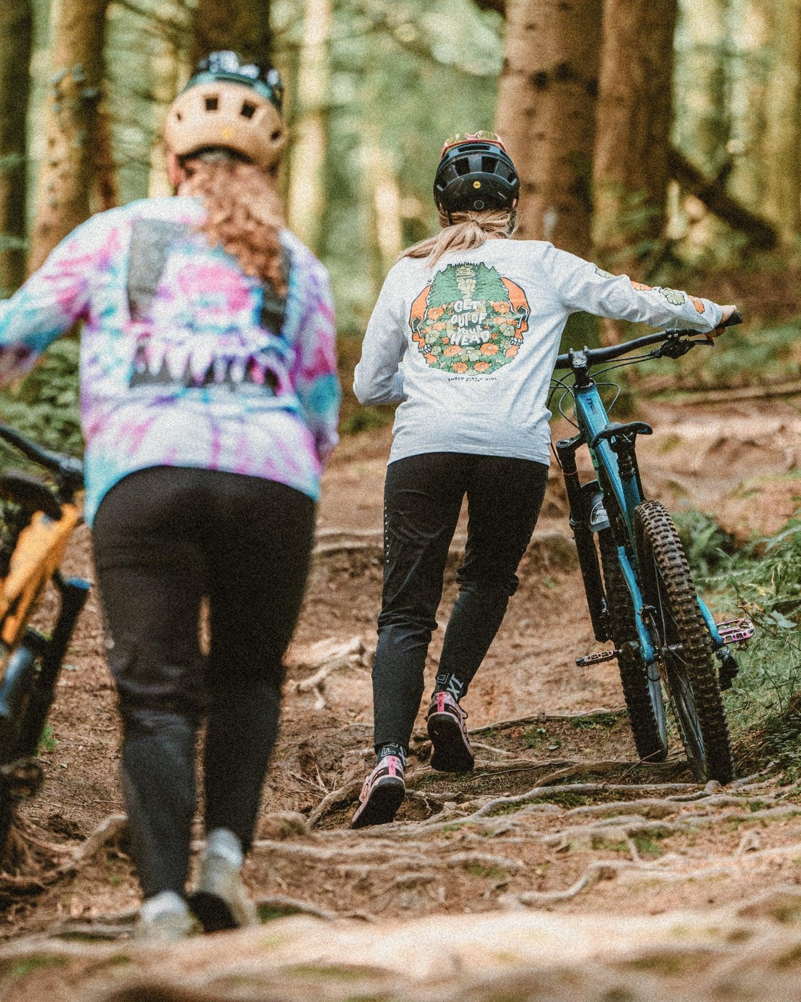 Two mountain bikers pushing bikes uphill on forest trail wearing colorful sustainable MTB apparel and helmets