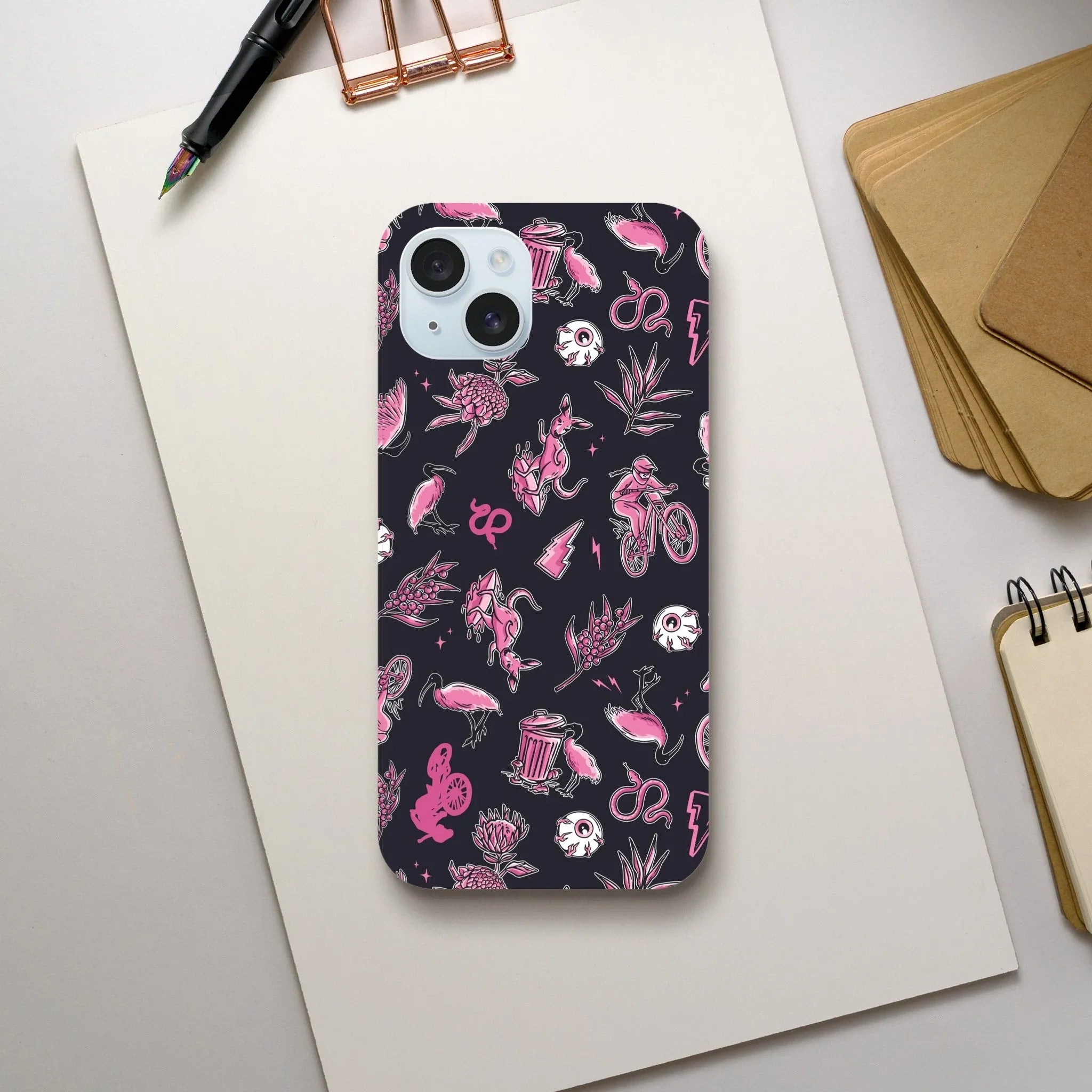 iPhone case with black background featuring pink Aussie-themed icons including kangaroo, cyclist, flamingo, flowers, and skull on white desk